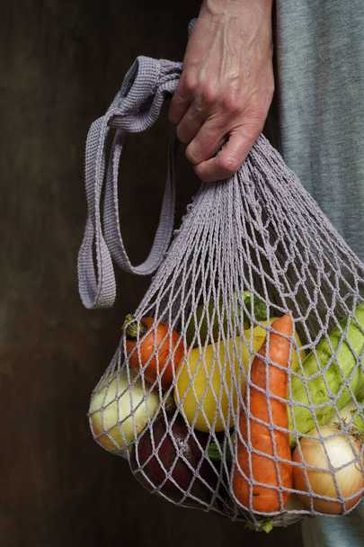 Woman holds her hand gray bag string bag with vegetables,zucchini,bell peppers,carrots,beets,onions brown wooden background.Harvest from garden fresh bright vegetables.Concept zero waste. - Фото, изображение