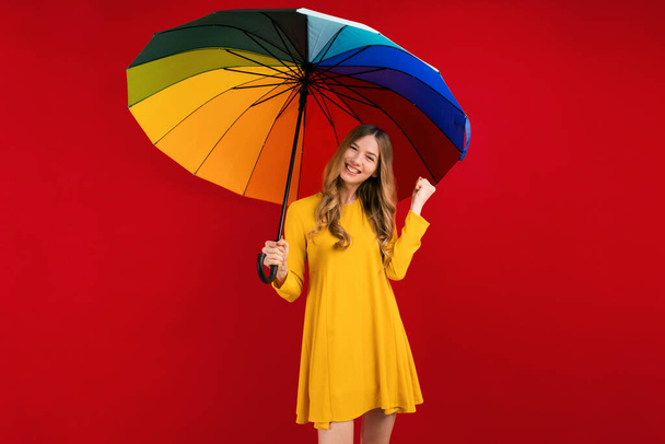 Happy young woman with a rainbow umbrella, shows a winning gesture on a red background, autumn concept - Fotoğraf, Görsel