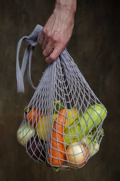 Vrouw houdt haar hand grijze zak string tas met groenten, courgette, paprika 's, wortelen, bieten, uien bruin houten achtergrond.Oogst uit de tuin verse heldere groenten.Concept nul afval. - Foto, afbeelding