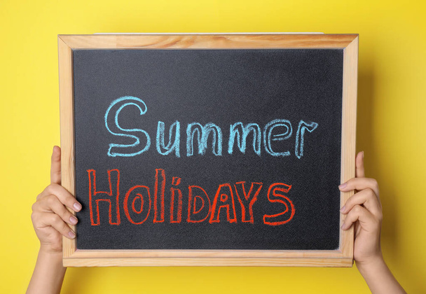 Schoolgirl holding blackboard with text SUMMER HOLIDAYS on yellow background, closeup - Foto, afbeelding