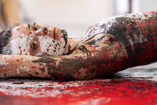 junge und sexy Frau in Unterwäsche, Sportbekleidung, künstlerisch abstrakt bemalt mit weißer, roter und schwarzer Farbe, liegend auf dem bunt bemalten Boden im Künstleratelier, Kopierraum. - Foto, Bild