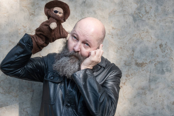 Portrait of a idiosyncratic crazy artist puppeteer with beard, holding a robber doll in front of his eye, copy space - Photo, Image