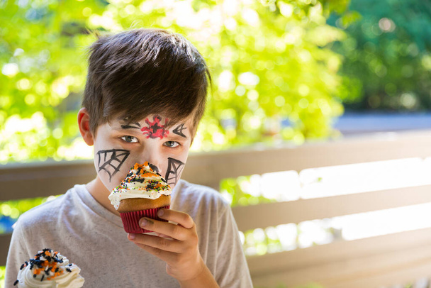 Niño con una cara de pintura comiendo espeluznantes cupcakes de Halloween - Foto, Imagen