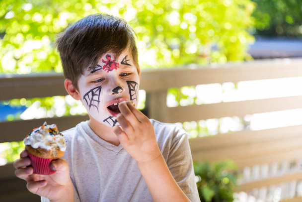 Menino com uma pintura facial comendo cupcakes assustadores de Halloween - Foto, Imagem