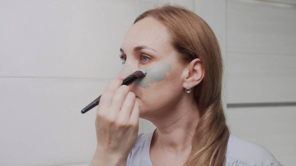 Κοντινό πλάνο της ενήλικης γυναίκας που φορά πήλινη μάσκα στο πρόσωπό της κοιτάζοντας στον καθρέφτη - Φωτογραφία, εικόνα