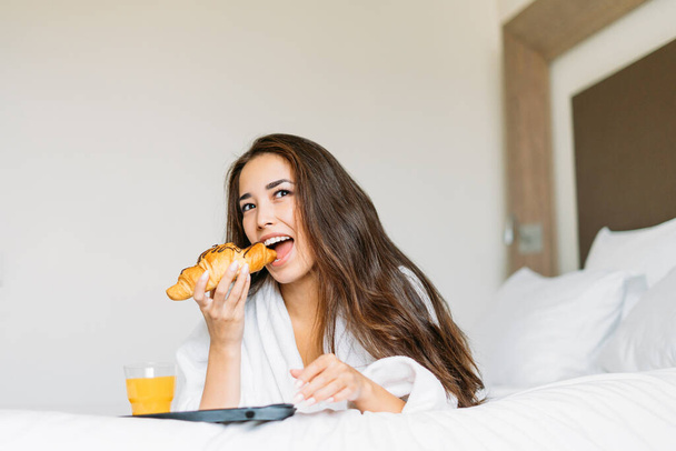 Beautiful young asian woman with long hair in white robe having breakfast croissant and orange juice in bed of hotel room - Foto, Imagen