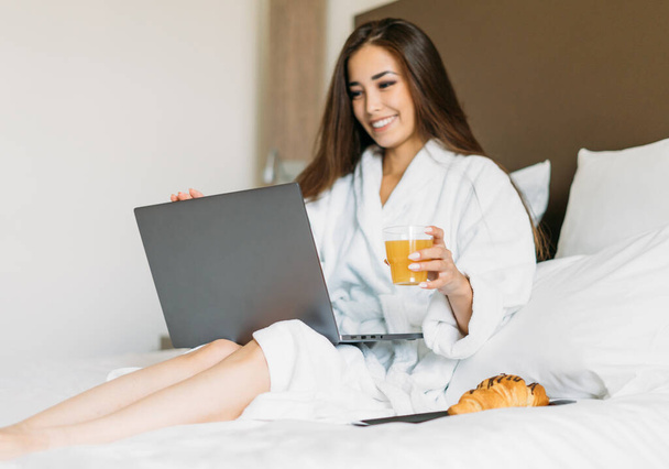 Młoda azjatycka dziewczyna z długimi włosami w białej szacie przy użyciu laptopa jedząc śniadanie croissant i sok pomarańczowy w łóżku w pokoju hotelowym - Zdjęcie, obraz