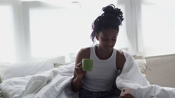 Close up of Young African American Woman uses phone while Relaxing in Bed - Foto, Imagem