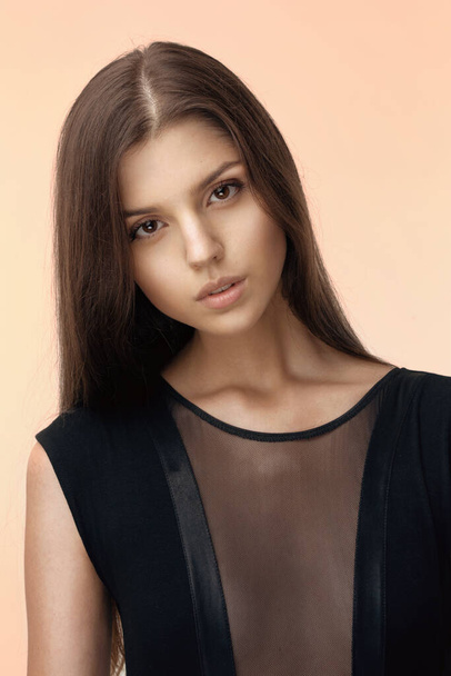 Portrait of young beautiful brunette girl in sensual black dress, looking at camera over beige background. - 写真・画像