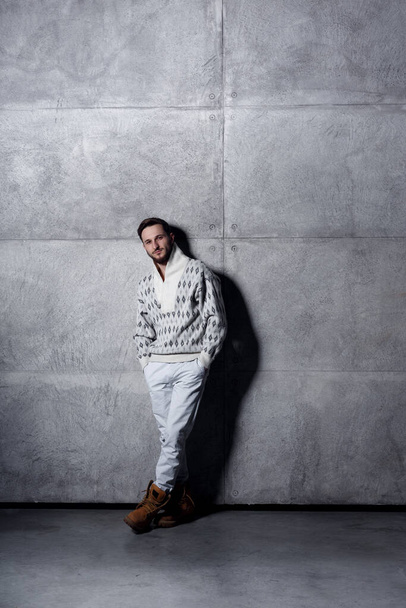 Vertical view of a guy in white jeans and sweater, posing in studio near concrete wall background. - Foto, afbeelding