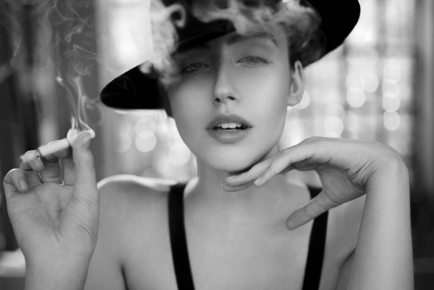 Schwarz-Weiß-Bild einer rauchenden jungen Frau mit schwarzem Hut, auf einem Stuhl sitzend, ihr Kinn zart mit der Hand berührend, isoliert in einem verschwommenen Innenraum, hinter einem Fenster. - Foto, Bild