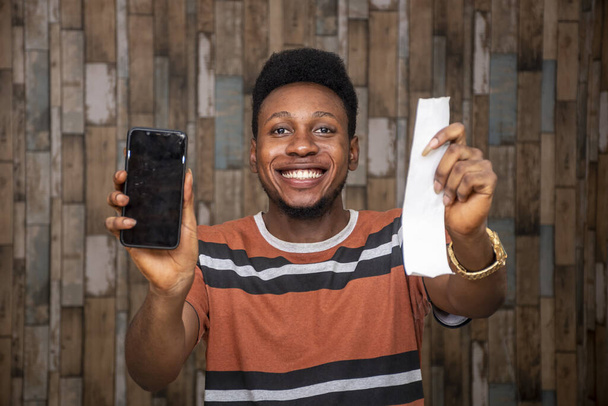 A young African male feeling excited while holding his smartphone and a paper slip - Foto, imagen
