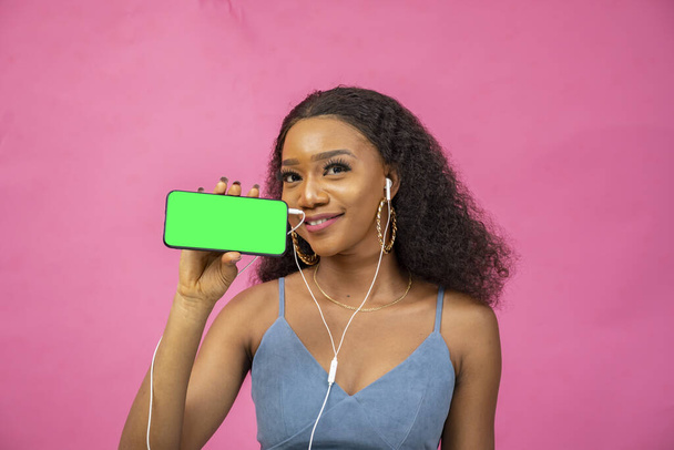 A young African woman wearing headphones while showing her phone screen on a pink background - Foto, immagini