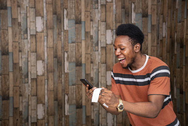 A young African male feeling excited while holding his smartphone and a paper slip - Photo, image