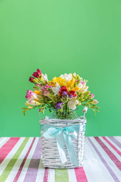 Boeket bloemen in een glazen vaas geplaatst in een rieten houten container waarop een lichtblauw lint is geplaatst. De vaas wordt geplaatst op een tafel bedekt met een veelkleurig tafelkleed tegen een groene achtergrond. - Foto, afbeelding