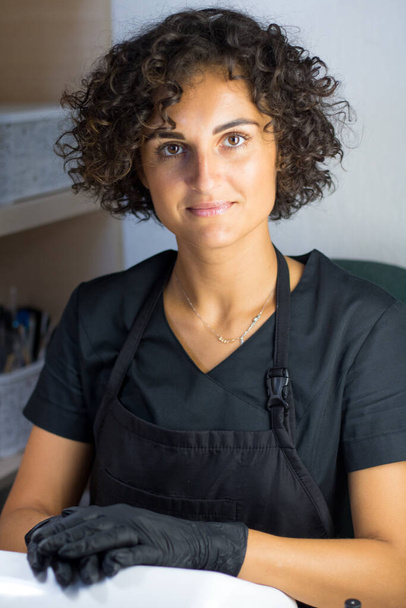 junge kaukasische Maniküre-Meisterin in schwarzen Handschuhen lächelt am Arbeitsplatz - Foto, Bild