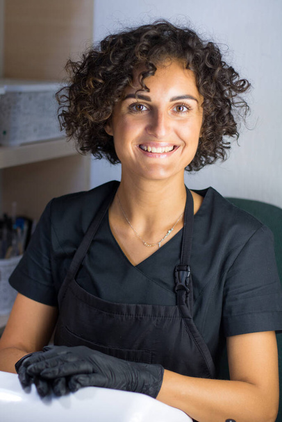 young caucasian woman manicurist master in black gloves is smiling on the working place - Photo, Image