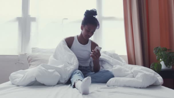 かなり若いアフリカ系アメリカ人女性はベッドでリラックスしながら電話を使用します - 映像、動画