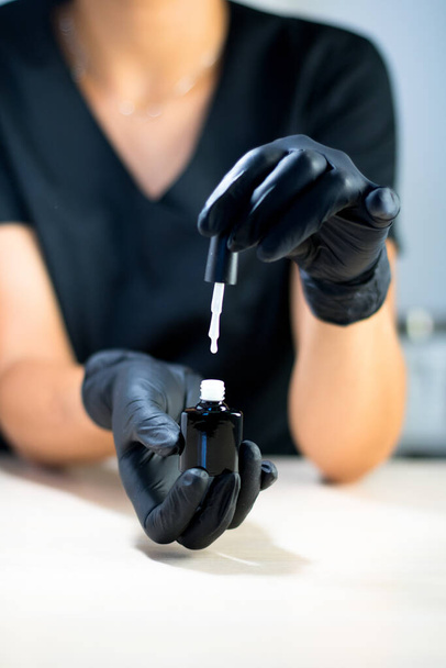 Мастер маникюра в черных перчатках держит белый лак для ногтей над столом - Фото, изображение