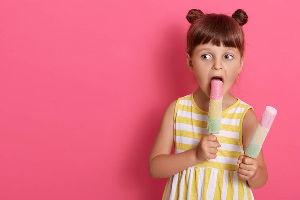 Little cute girl with widely opened mouth biting water ice cream, looking aside while posing isolated over pink background, copy space for advertisement. - Zdjęcie, obraz