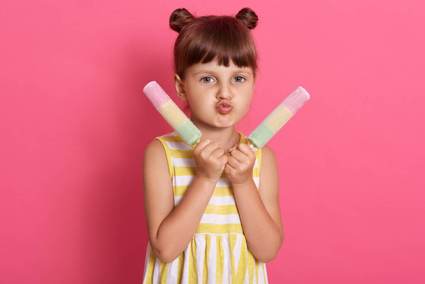 Girl with two water ice cream looking at camera and making grimace, pout lips, wearing striped summer dress, standing isolated over pink background. - Фото, зображення