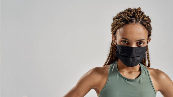 Coronavirus and sport. Confident sportive mixed race woman wearing black medical protective face mask looking at camera while standing isolated over grey background - Fotografie, Obrázek