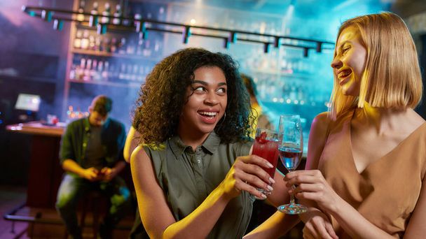 Attractive young women looking at each other while toasting, posing with cocktail in their hands. Friends celebrating, having fun in the bar - Foto, Imagem