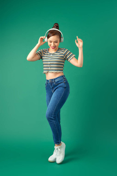 Full length portrait of joyful Asian woman on casual clothing dancing and listening to music with headphone over green background. - 写真・画像