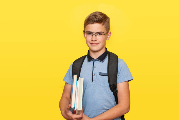 A schoolboy teenager with glasses with books in his hands and a school backpack smiles against a yellow background. Back to school. - Foto, Imagem