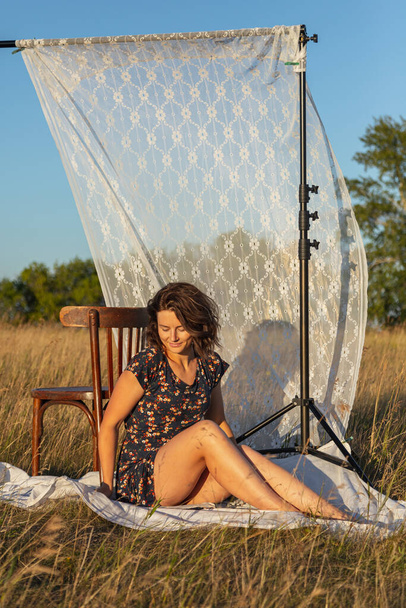 Egy gyönyörű, virágos ruhás fiatal nő portréja, aki egy fa széken ül egy fehér csipke függöny hátterében a pályán egy napos nyári napon..  - Fotó, kép