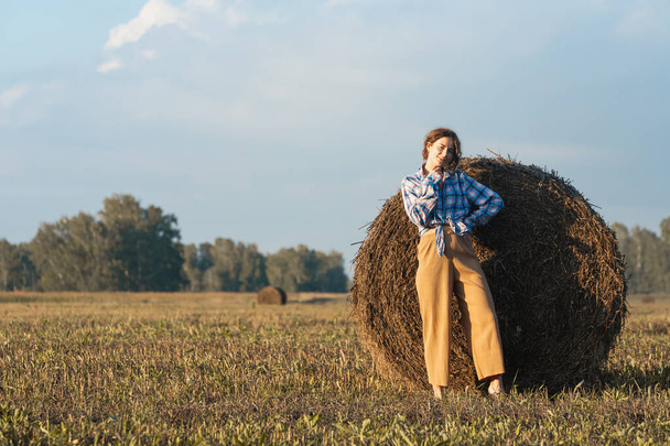 Hermosa mujer joven moderna con pantalones beige y una camisa a cuadros posando contra un pajar en el campo. Chica adolescente de moda en otoño al aire libre.  - Foto, imagen