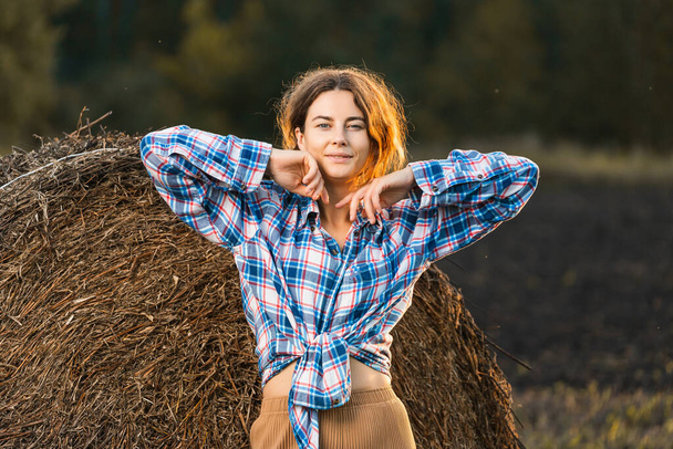 Portrait of a beautiful young model in beige trousers and a plaid shirt  have fun,  posing against on haystacks in  sunny autumn day . Autumn warm photo. Woman smiling and look away, joyful cheerful mood. - Foto, afbeelding