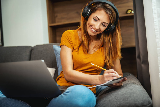 Cute young female student sitting on the sofa, using her laptop and headphones to watching lesson online and learning from home. Young woman taking notes while following teacher doing lesson on video call.  - Φωτογραφία, εικόνα