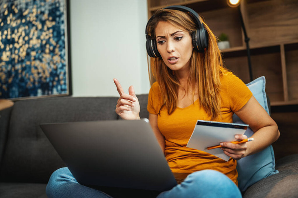 Cute young female student sitting on the sofa, using her laptop and headphones to watching lesson online and learning from home. Young woman taking notes while following teacher doing lesson on video call.  - Fotoğraf, Görsel