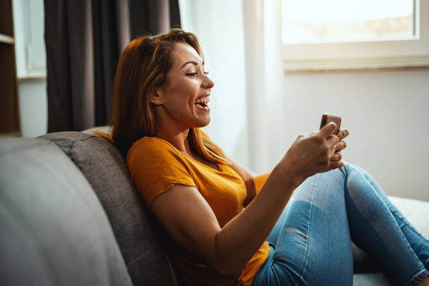 Aantrekkelijke jonge vrouw zitten op de bank en het gebruik van de smartphone om videospel te spelen thuis. - Foto, afbeelding