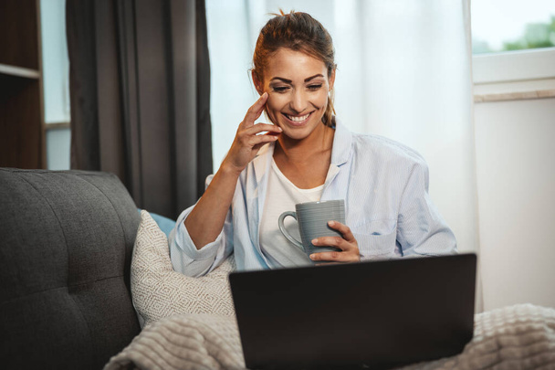 Mulher bonita sentada no sofá, desfrutando de uma xícara de café e usando seu laptop em casa. - Foto, Imagem
