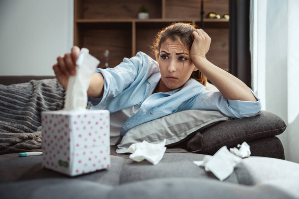 Young woman is lying sick at home couch and taking paper handkerchief for blowing nose. - Foto, Imagen