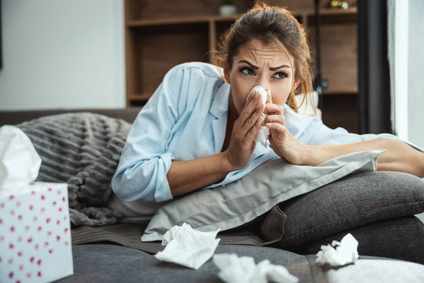 Νεαρή γυναίκα είναι άρρωστη στο σπίτι καναπέ φυσάει μύτη με μαντήλι. - Φωτογραφία, εικόνα