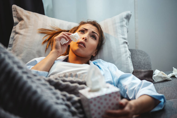 Молода жінка лежить хворий вдома диван з паперовою носовичкою в руці за дуючий ніс
. - Фото, зображення