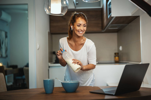 Beautiful young woman is preparing her healthy breakfast in her kitchen and using laptop to make a video chat with someone. - Photo, image