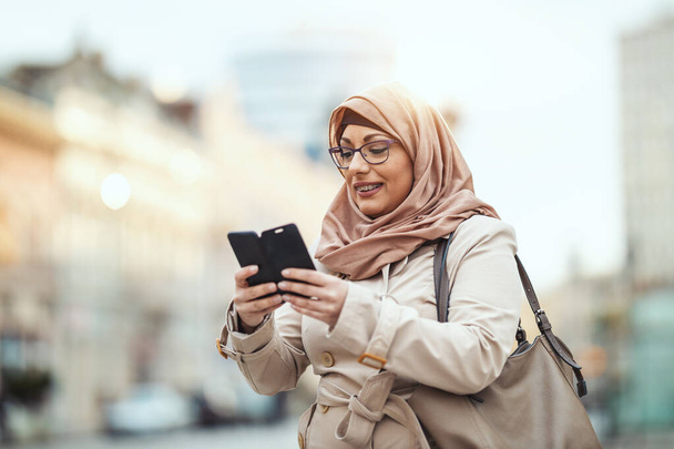 Moslim vrouw van middelbare leeftijd draagt hijab met een vrolijk gezicht in stedelijke omgeving, tippen berichten op haar smartphone. - Foto, afbeelding