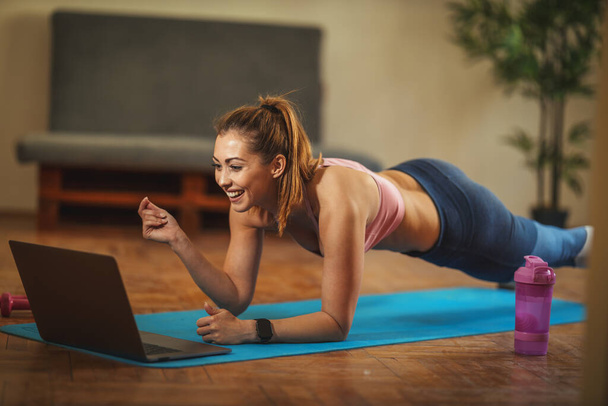 Young smiling woman is doing plank exercises in the living room on floor mat at home, looking at the laptop, in morning sunshine. - Фото, зображення