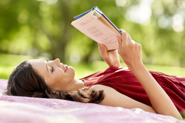 happy woman reading book lying on blanket at park - Photo, Image
