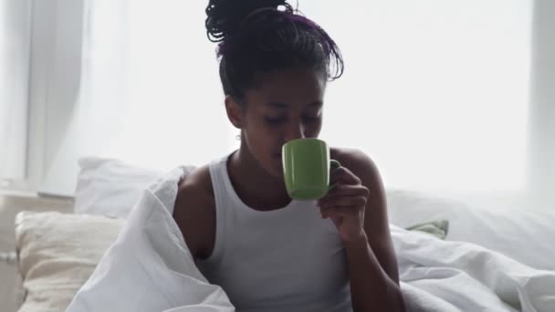Close up de jovem afro-americana mulher senta-se na cama, bebe café da manhã e usa telefone - Filmagem, Vídeo