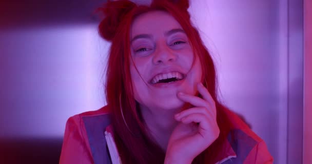 Close up of charming female person looking to camera and laughing. Portrait of happy young woman with broad smile touching her face with hand while possing in neon lights. - Filmati, video