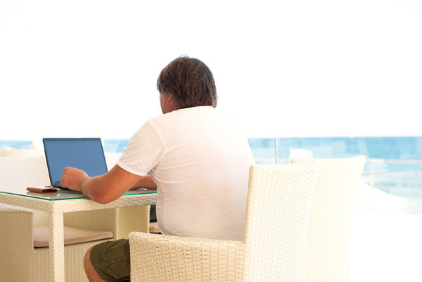 Miejsce pracy w kawiarni z widokiem na morze. Facet pracujący nad laptopem w kawiarni. Przestrzeń kopiowania, Mock up. - Zdjęcie, obraz