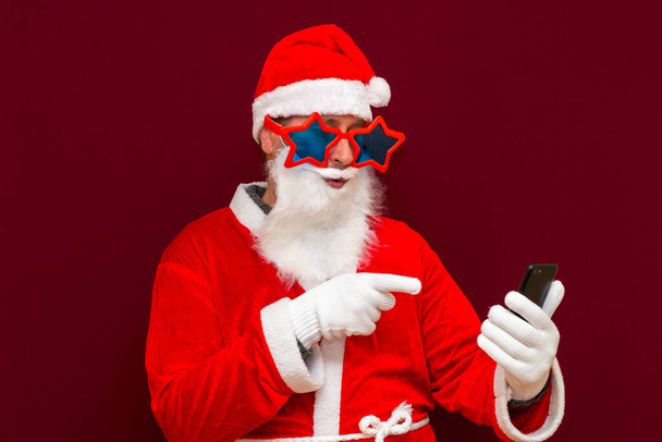 Retrato de moda Santa Claus utilizar teléfono celular comentario entradas de año nuevo con estilo estrellas de moda gafas gafas sombrero leer noticias aisladas sobre fondo rojo - Foto, Imagen