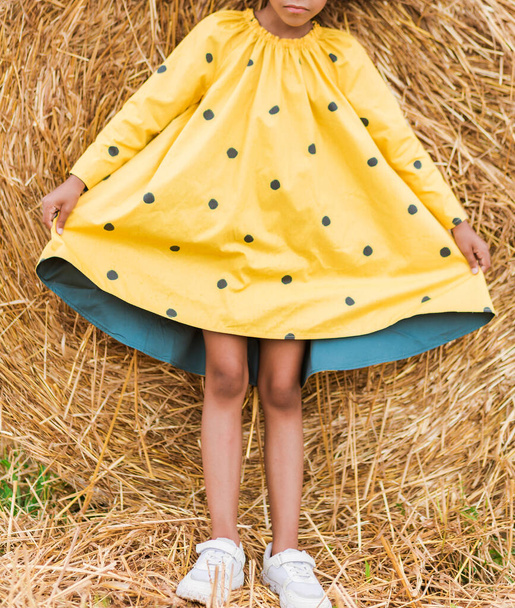 Little african american girl in a trendy yellow dress with polka dots and white athletic shoes posing against the background of a haystack. Vertical photo - Fotoğraf, Görsel