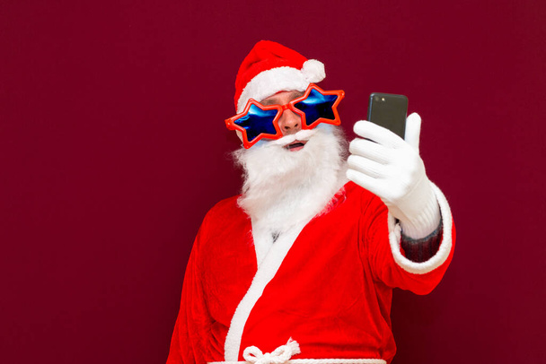 Portrait of trendy Santa claus use cell phone comment newyear posts wearing stylish trendy stars eyewear eyeglasses hat reading news isolated over red background - Photo, Image