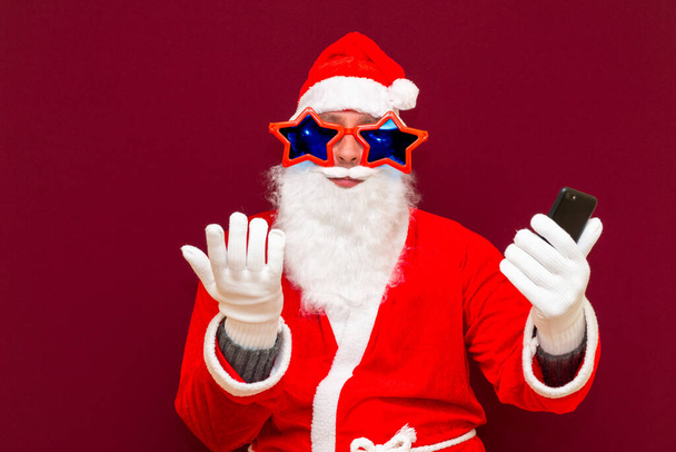 Portrét módní Santa Claus použít mobilní telefon, co se stalo. Nosí stylové trendy hvězdy brýle klobouk čtení zprávy izolované přes červené pozadí - Fotografie, Obrázek